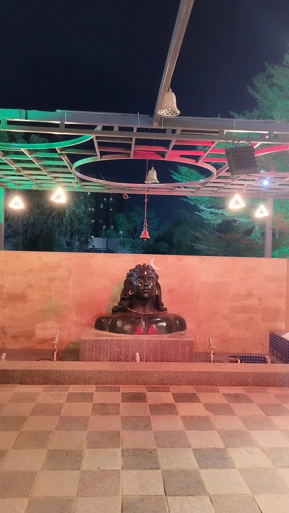 Black stone statue of Lord Shiva in an outdoor temple setting with hanging bells and decorative lighting at night.