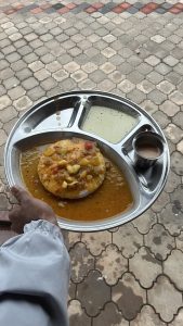 A person holds a metal plate containing a round, soft dish topped with a mix of diced vegetables and a rich, yellow-brown sauce.