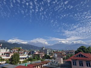 A panoramic view of a city with multi-story buildings and houses set against a backdrop of majestic snow-capped mountains under a clear blue sky decorated with wispy clouds