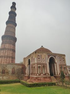 A tall, intricately designed minaret stands prominently on the left side of the image, showcasing its red and white stonework and detailed carvings. Adjacent to it is a smaller, ornate structure with a domed roof and decorative elements along its facade