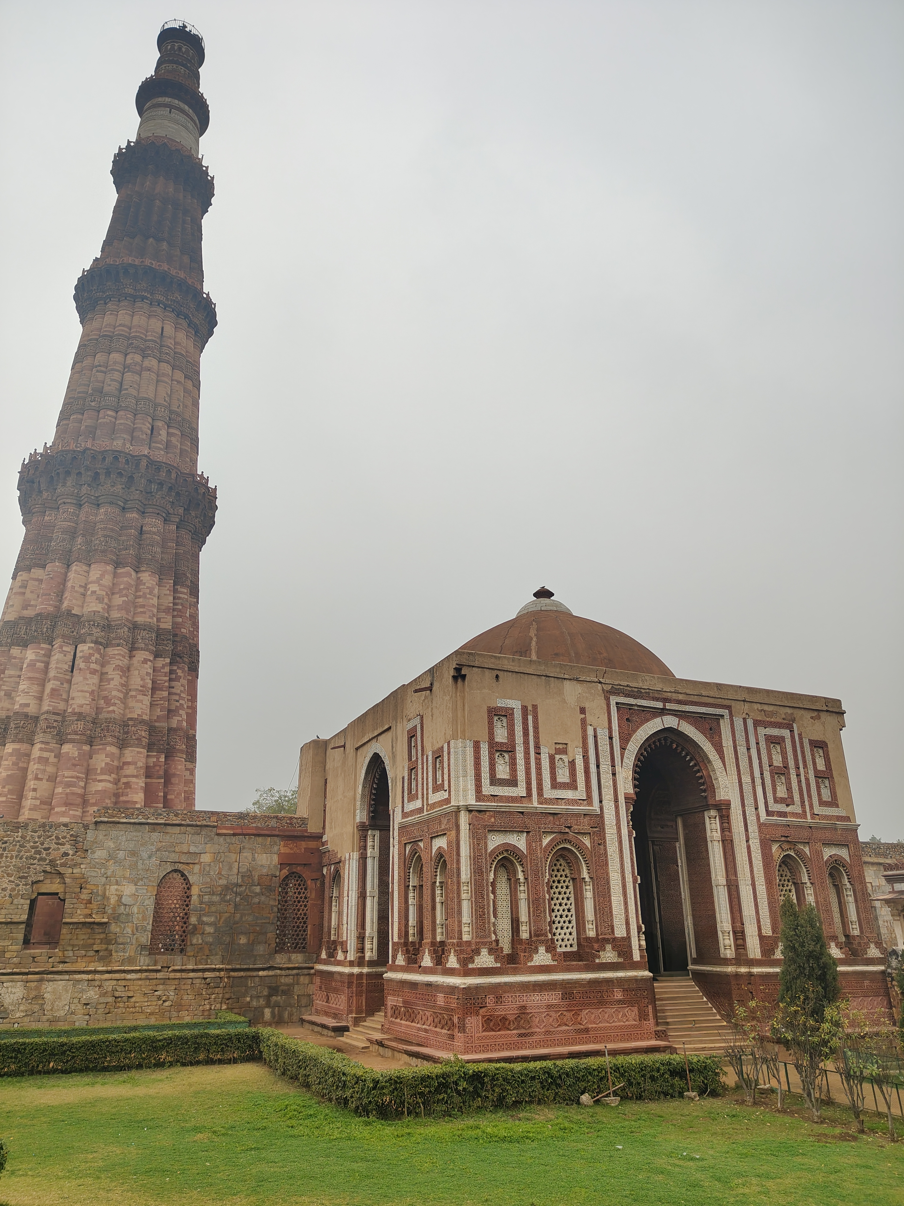 A tall, intricately designed minaret stands prominently on the left side of the image, showcasing its red and white stonework and detailed carvings. Adjacent to it is a smaller, ornate structure with a domed roof and decorative elements along its facade