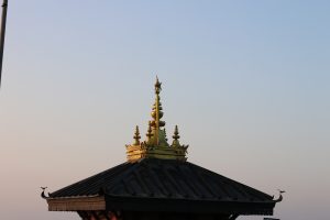 An ornate traditional roof with a golden carved apex and spires against a clear blue sky, with a small bird perched on the edge.