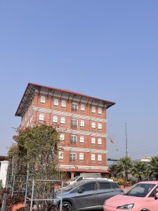 A red brick building with a flat roof and white window frames stands against a clear blue sky. In front of the building are several parked cars, including a pink one, along with greenery and decorative plants.