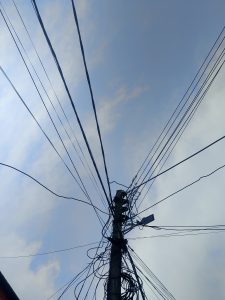 A view looking up at a utility pole with numerous tangled wires extending in multiple directions against a backdrop of a cloudy sky. 
