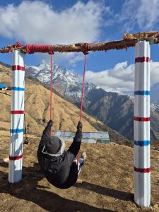 A person is swinging on a rustic swing set made of a wooden beam supported by two painted poles, with red and blue stripes.