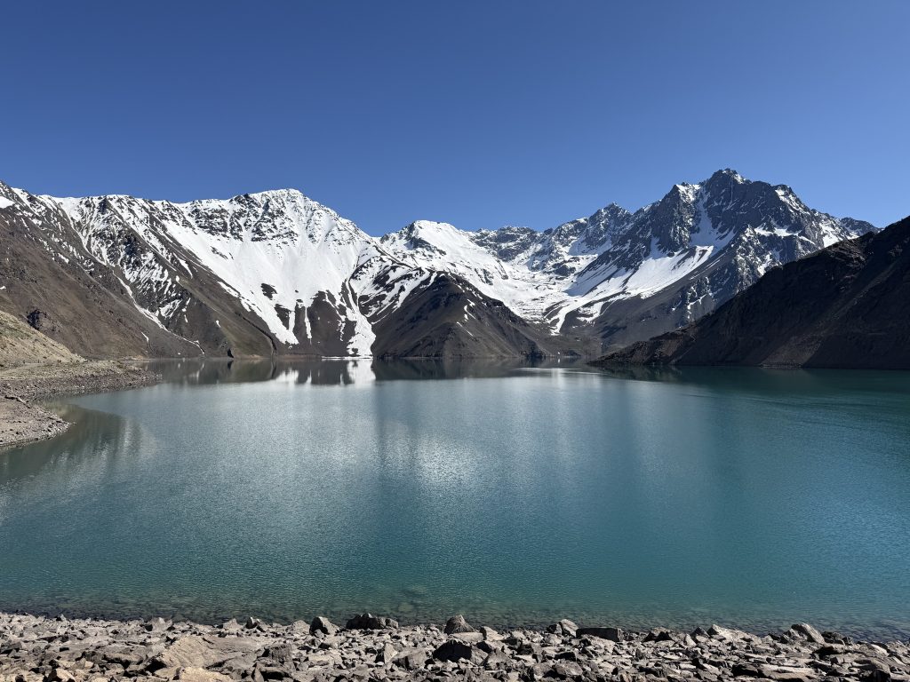 El Yeso Reservoir, Maipo Canyon, Chile, with a snow-patched peaks under a clear blue sky, with rocky ground in the foreground.
