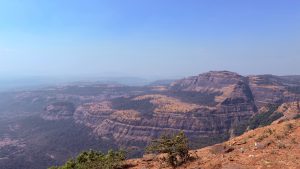 A panoramic view of Tiger Point, Lonavala with rugged mountains and rolling hills under a clear blue sky. The landscape features steep cliffs and layered rock formations, with patches of green vegetation interspersed among areas of brown earth. 
