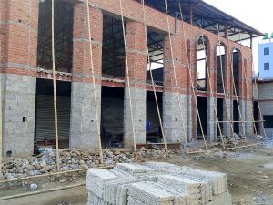 A brick building in the process of being built. There are large openings on the sides for future windows. There are wooden struts holding things up. There is a pile of bricks in the foreground.