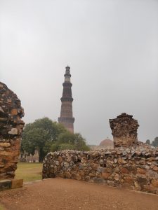 A tall ancient tower stands prominently in the background against a cloudy sky, surrounded by lush greenery. In the foreground, there are remnants of stone walls, showcasing a mix of rustic stones and disintegrating structures, suggesting historical significance