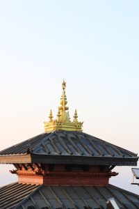 An ornate golden roof pinnacle with intricate details, set atop gray tiled roofing and a red-brick base against a soft pastel sky.