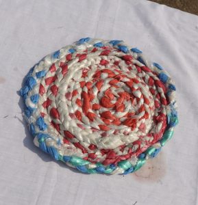 A circular mat made of braided plastic strips in a spiral pattern, featuring alternating colors of red, blue, and white, resting on a plain white surface.