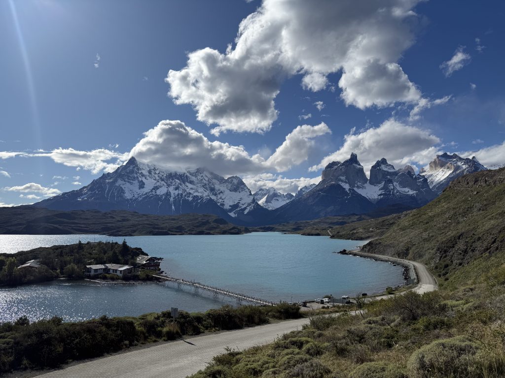 A calm turquoise lake with snow-capped mountains and green surroundings in Torres del Paine National Park, Chile.