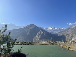 A scenic view of Dhumba Lake, a green-hued lake nestled in a mountain valley with snow-capped peaks in the background under a clear blue sky. 