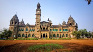 Seen from the front, a grand palace fills the view, with a clock tower rising from its center. In front of the palace is open land with some newly planted grass. Above it all, a clear blue sky adds to the peaceful scene.