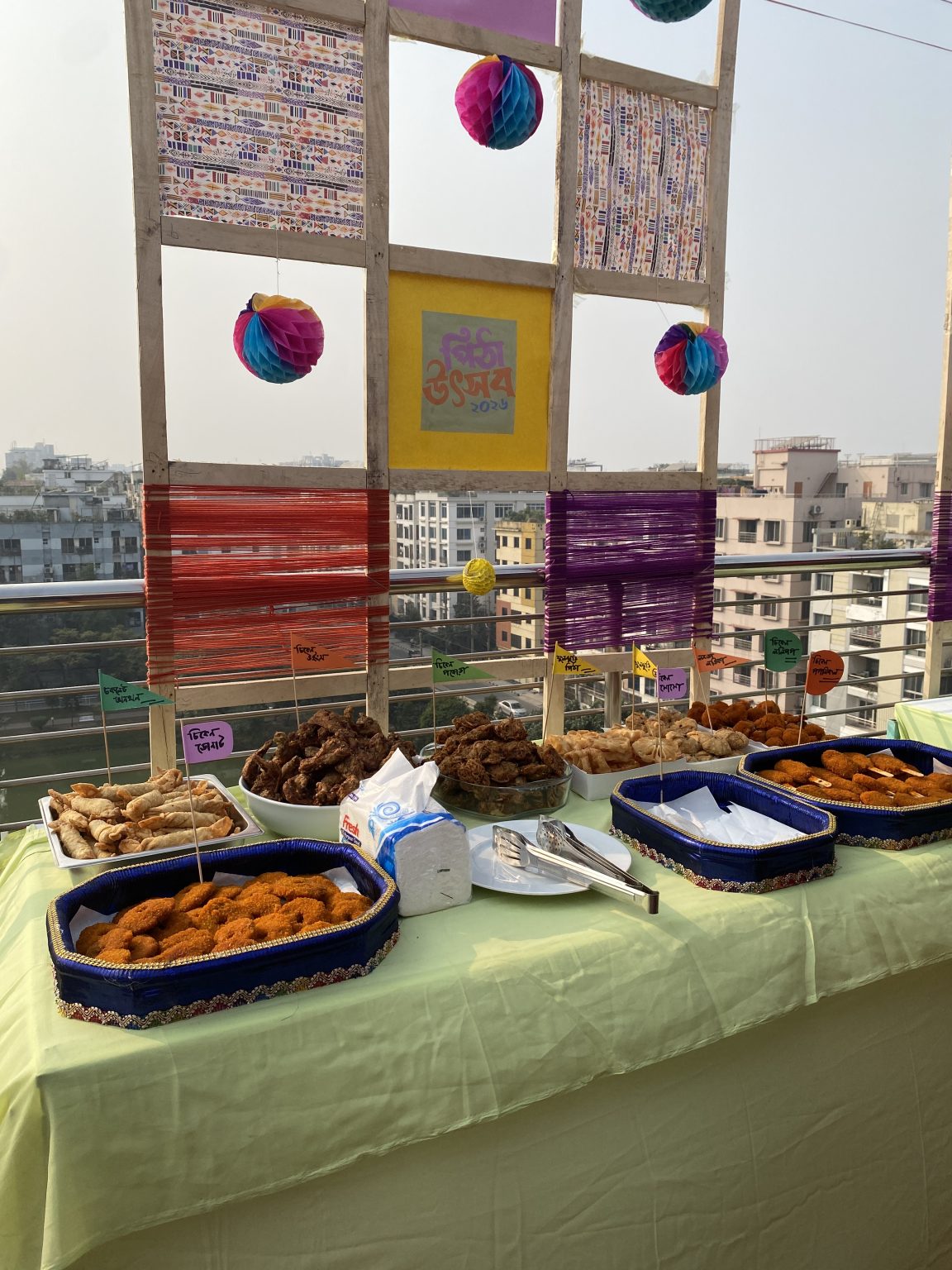 Pitha Festive, a Bengali Community celebration. A vibrant outdoor display of various fried snacks is presented on a table covered with a light green cloth. The snacks are arranged in blue and white trays, labeled with colorful signs indicating different names.