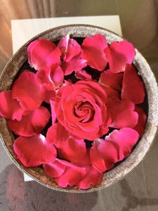
A round, decorative container filled with vibrant pink rose petals, with a single rose at the center. 