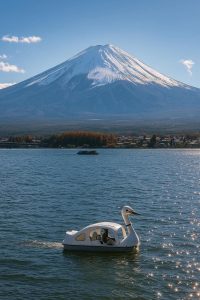 A calm lake with a white swan-shaped pedal boat on sparkling water. Snow-capped mountains rise in the background under a clear blue sky, with trees and small buildings along the shore.