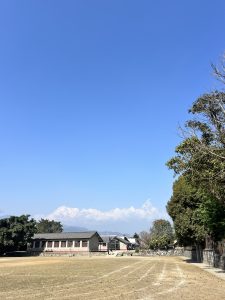 A calm scene with a grassy field, trees on one side, a building on the other, and snow-capped mountains under a clear blue sky.