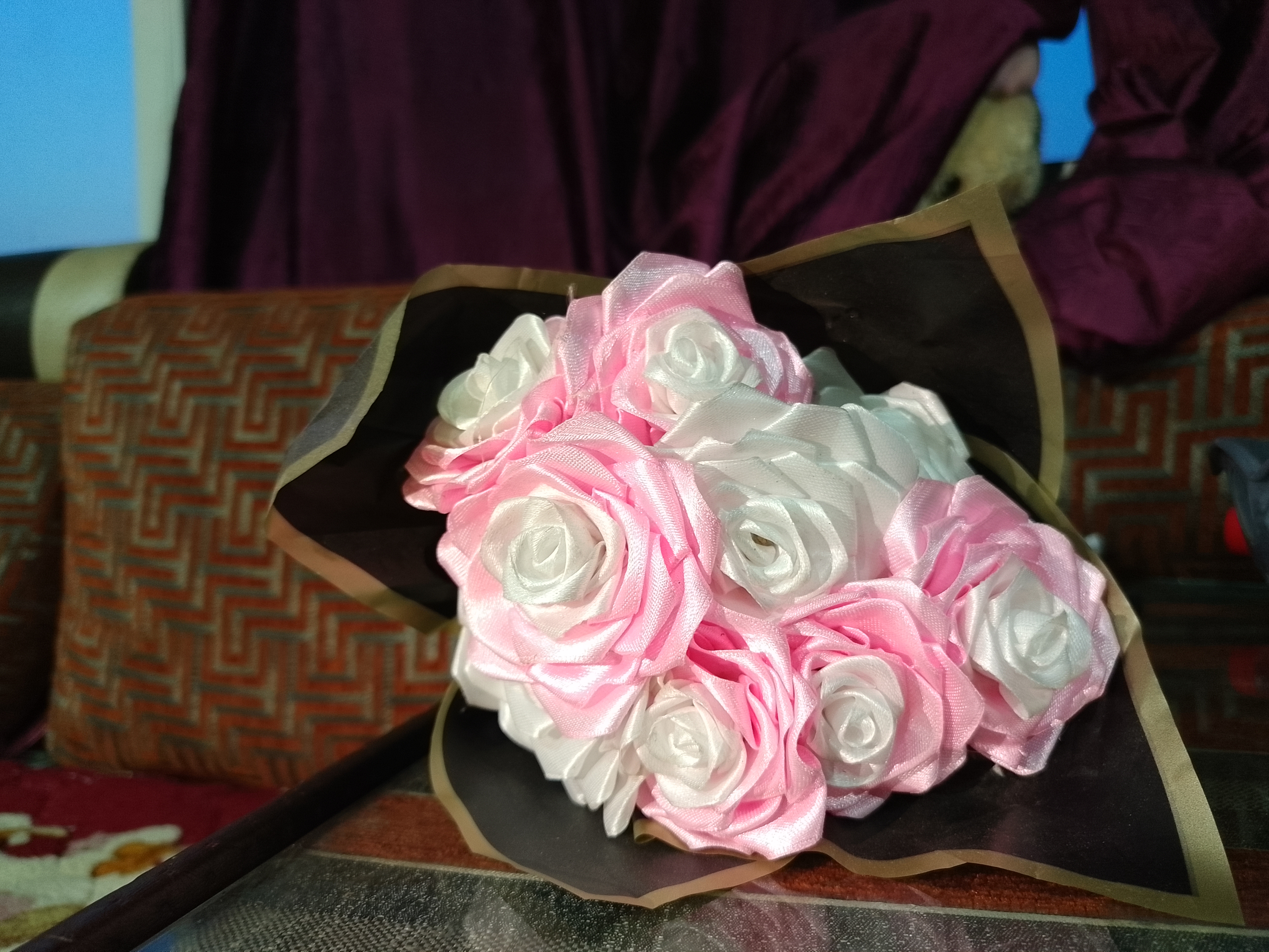 A bouquet of artificial roses, featuring a mix of soft pink and white flowers, is arranged beautifully. The roses are made from a satin-like material, giving them a shiny, smooth appearance. The bouquet is wrapped in dark brown paper with a subtle gold trim, adding an elegant touch.