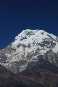 A snow-capped mountain peak rises prominently against a deep blue sky, showcasing a mix of rugged rock formations and gleaming ice. 
