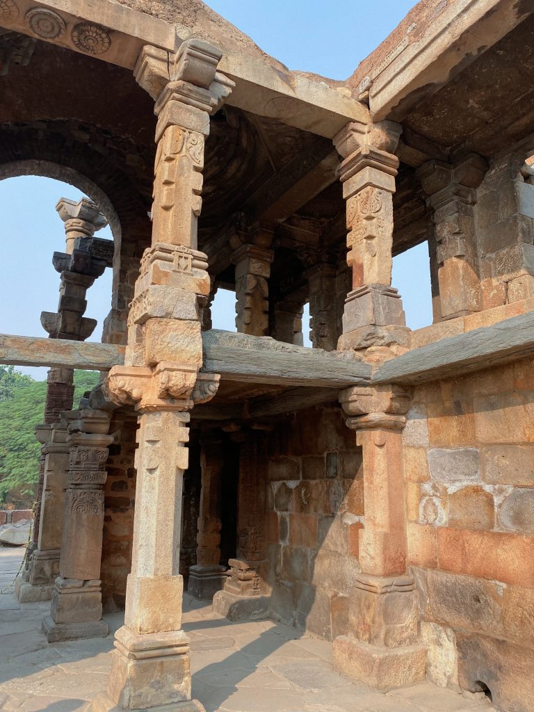 An ancient stone building with carved pillars and stone walls, partly roofed, with sunlight, shadows, and some greenery around.