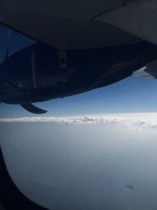 A view from an airplane window showing a blue and white airplane wing and propeller against a backdrop of fluffy white clouds and a clear blue sky. 