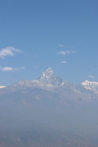 A distant view of a majestic fishtail (machapuchare) mountain peak, partially covered in snow, rising sharply against a clear blue sky.