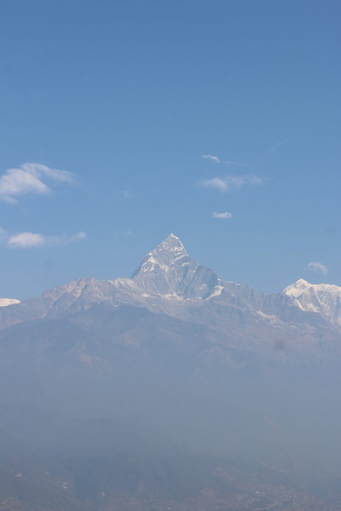 A distant view of a majestic fishtail (machapuchare) mountain peak, partially covered in snow, rising sharply against a clear blue sky.
