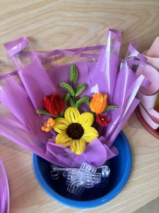 
A vibrant arrangement of artificial flowers, featuring a bright yellow sunflower and various colored flowers (red, orange, and green), is displayed in a blue bowl.