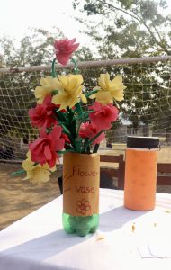 
A decorative flower vase made from a green plastic bottle, with a brown paper wrapper that reads "Flower vase" and includes a hand-drawn flower. 