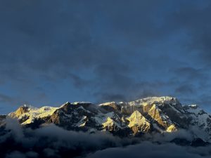 A dramatic mountain range covered in snow rises against a backdrop of a dark blue sky, with patches of golden light illuminating the peaks. 