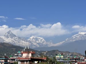 A panoramic view of snow-capped mountains under a clear blue sky, with some clouds scattered in the background.