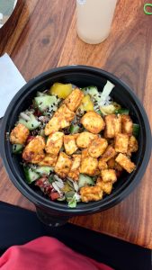 A black bowl filled with a colorful salad featuring pieces of spiced tofu, diced cucumbers, cherry tomatoes, yellow bell peppers, and various seeds
