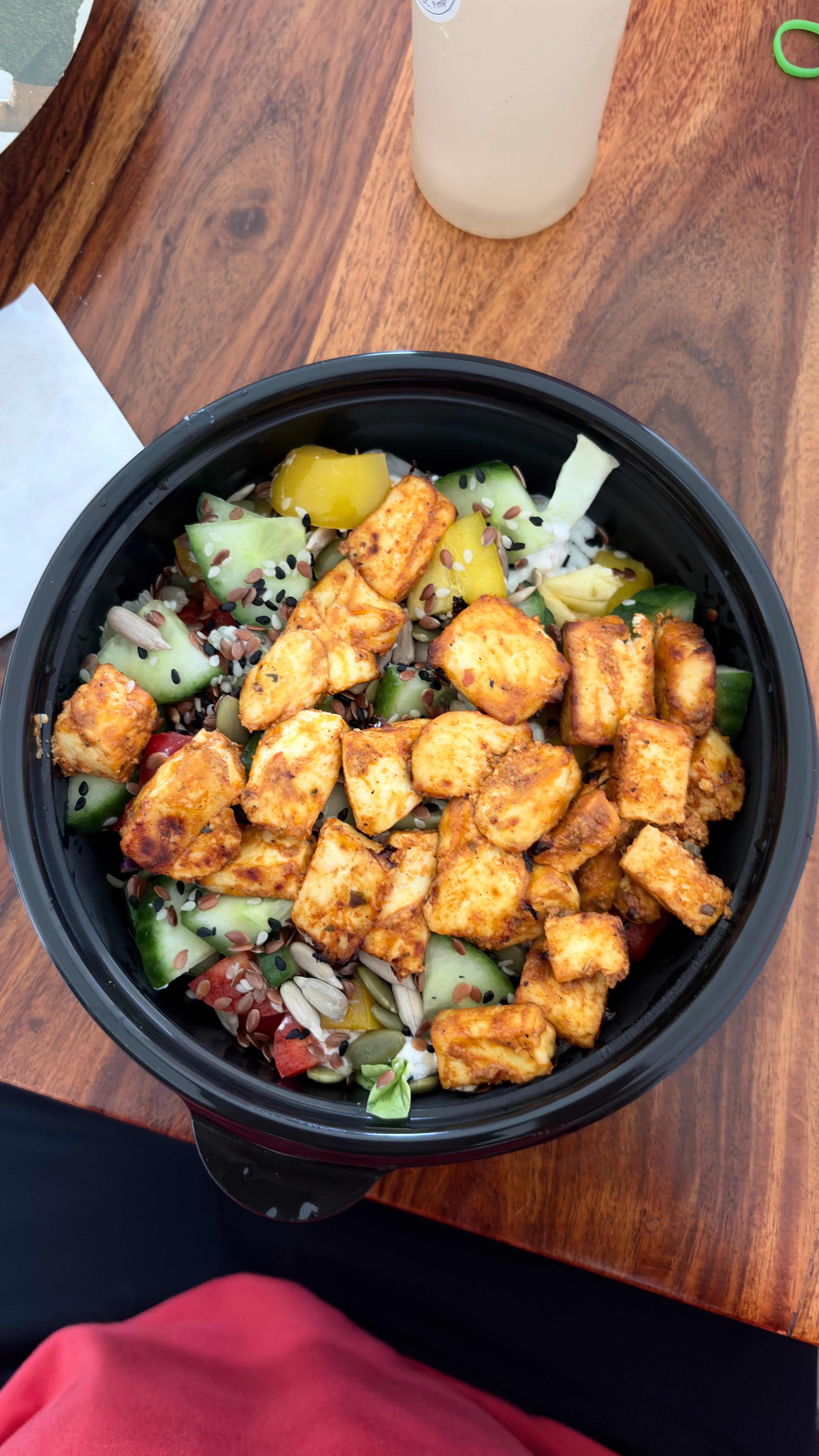 A black bowl filled with a colorful salad featuring pieces of spiced tofu, diced cucumbers, cherry tomatoes, yellow bell peppers, and various seeds