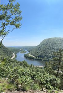 A panoramic view of a winding river surrounded by lush green hills under a clear blue sky. 
