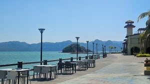 Seaside boardwalk with outdoor café tables and chairs overlooking turquoise water, small forested islands, and distant mountains under a clear blue sky.