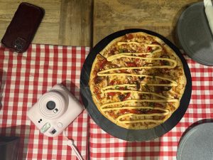 A round pizza with red peppers and creamy sauce sits on a black plate atop a red-and-white checkered tablecloth. Nearby are a pink instant camera, a dark phone case, a grey plate, and a napkin.