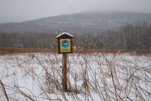 A yellow State Land Forest Preserve sign posted on a birdhouse on a post at the edge of a snowy field with mountain foothills in the background.
