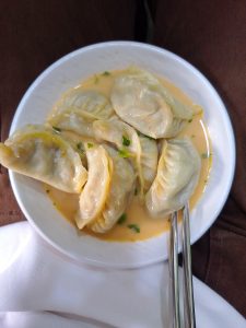 A white bowl filled with six dumplings in a creamy, light orange sauce, garnished with small pieces of green herbs.