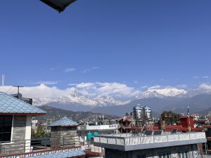 A wide view of snow-covered mountains under a blue sky. In front, there are buildings with different rooftops, some blue, along with water tanks and solar panels, showing a mix of city life and mountain scenery.
