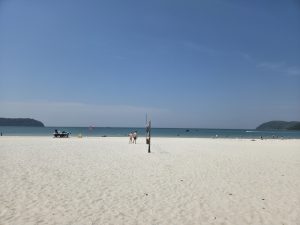 A sunny beach scene. A broad expanse of soft, white sand stretches across the foreground. In the background, a calm blue sea meets the horizon, dotted with a few boats.