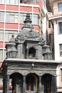 The top section of the ancient, multi-tiered stone temple has intricate carvings and a dark, weathered appearance. It is built with wood and located in front of tall buildings.
