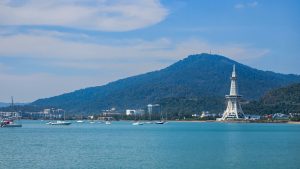 Coastal scene featuring a calm, turquoise bay dotted with various boats.In the background, a lush green mountain rises, partially covered with trees. On one side of the bay, there are modern buildings, tall tower with a lattice design stands prominently against the skyline.