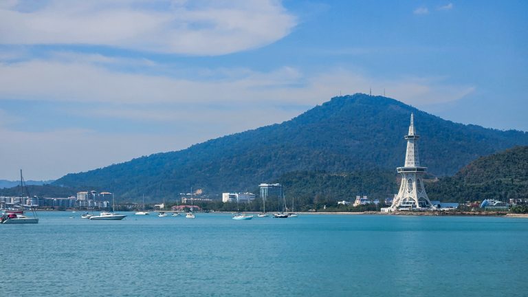 Coastal scene featuring a calm, turquoise bay dotted with various boats.In the background, a lush green mountain rises, partially covered with trees. On one side of the bay, there are modern buildings, tall tower with a lattice design stands prominently against the skyline.