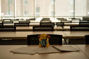 A room full of tables with a glass wall behind. The closest table is clear, but the rest out of focus. In the foreground is a small plush toy (wapuu), with a pen leaning on it, with papers and notebooks arrayed in front of it on the table.