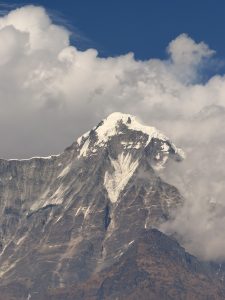 
A snow-capped mountain peak rises above a rugged landscape, partially obscured by white clouds. 
