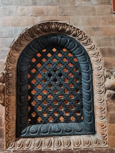 An intricately carved window frame featuring a lattice design with diamond shapes and floral motifs around the arch.