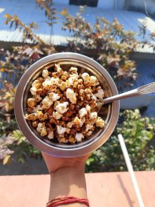 A hand holding a bowl of popcorn, including both white and caramel-coated kernels, with a silver spoon resting on the side