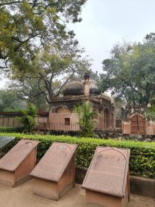 A historical structure features two large domes and ornate architectural details, surrounded by greenery and trees. In the foreground, there are three informational plaques with text, placed on a grassy area near a low hedge.