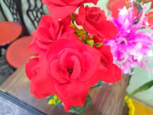 
A close-up view of a bouquet of artificial red roses, showcasing their bright, vibrant petals. In the background, there are hints of other flowers and round, red-topped chairs